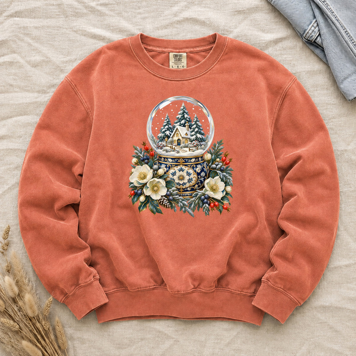Snow Globe & Winter Florals Sweatshirt in Terracotta — a glass snow globe holding a snowy cottage and evergreens on an ornate navy-and-gold base, framed by cream blossoms, pinecones and red berries