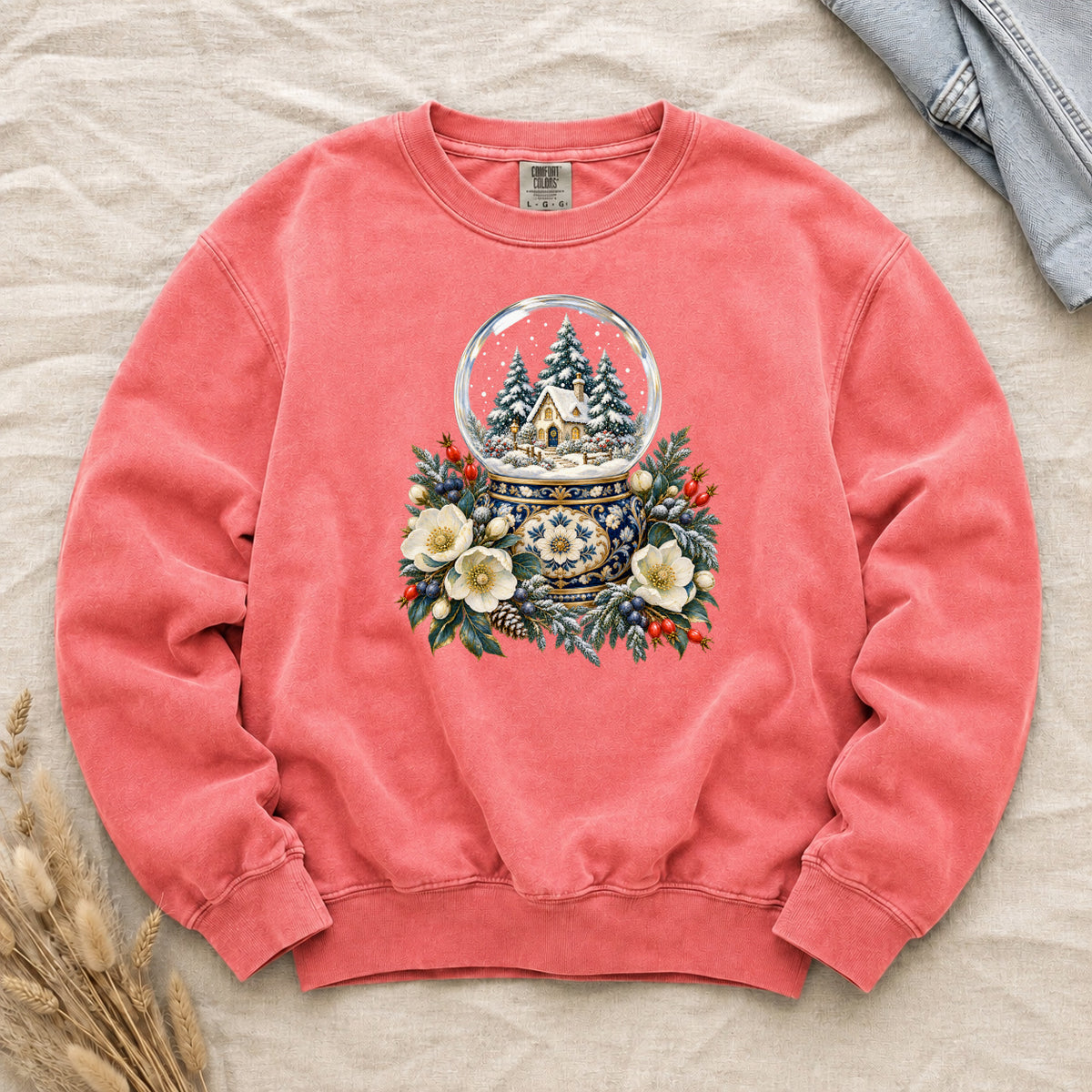 Snow Globe & Winter Florals Sweatshirt in Watermelon — a glass snow globe holding a snowy cottage and evergreens on an ornate navy-and-gold base, framed by cream blossoms, pinecones and red berries