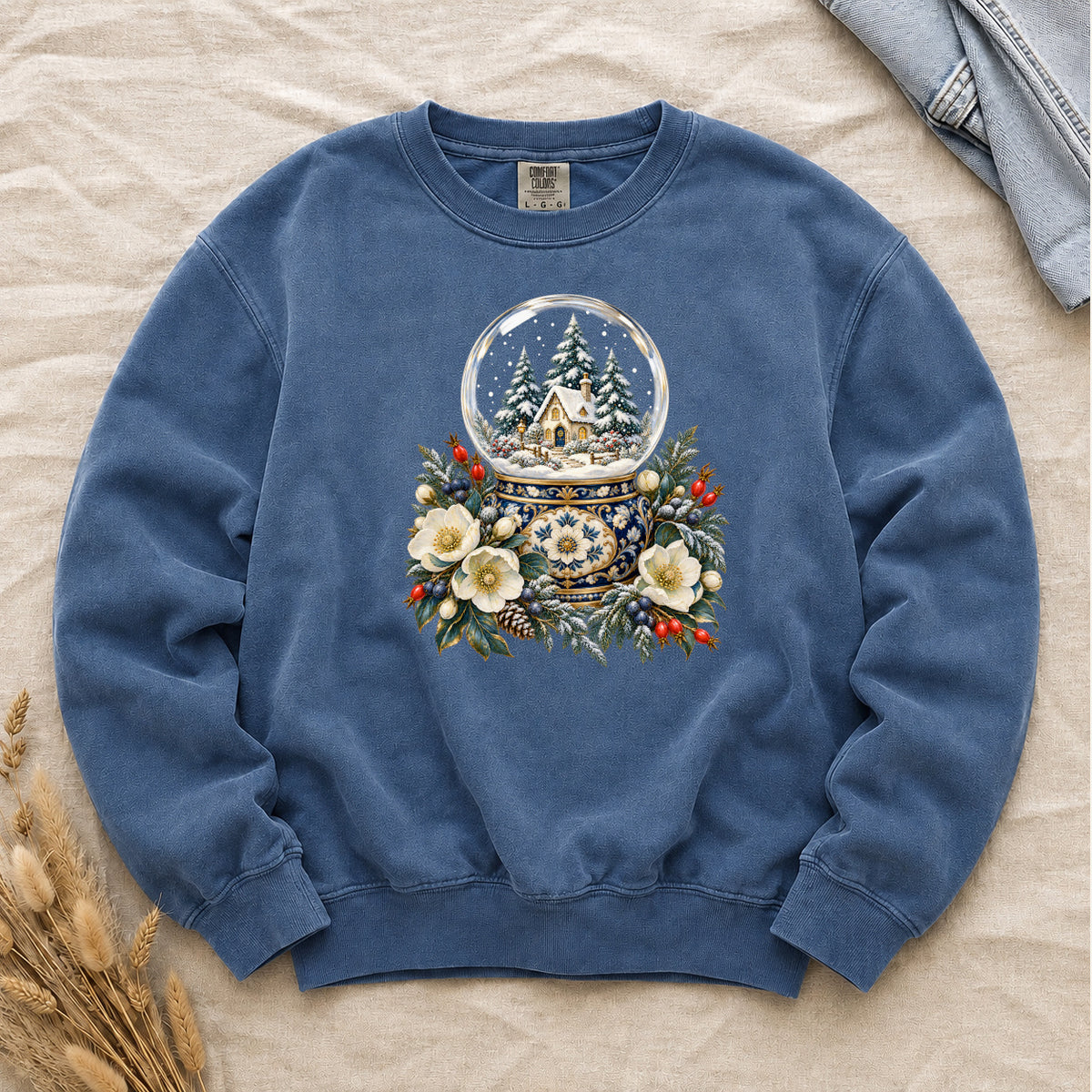 Snow Globe & Winter Florals Sweatshirt in Blue Jean — a glass snow globe holding a snowy cottage and evergreens on an ornate navy-and-gold base, framed by cream blossoms, pinecones and red berries
