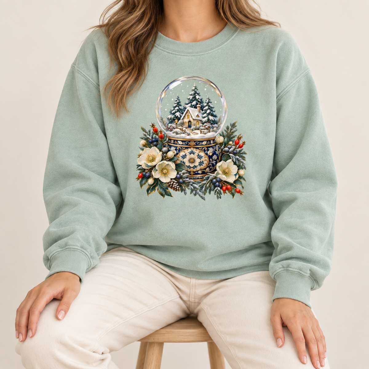 Vintage holiday snow globe sweatshirt in heavyweight garment-dyed cotton — a glass snow globe holding a snowy cottage and evergreens on an ornate navy-and-gold base, framed by cream blossoms and red berries