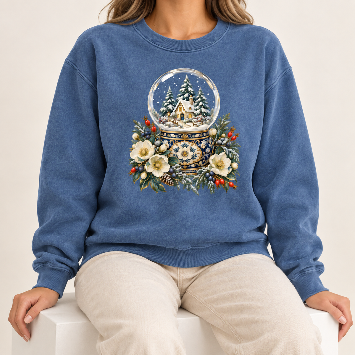 Snow globe and winter botanical design on an 80/20 cotton-polyester crewneck — a glass snow globe holding a snowy cottage and evergreens on an ornate navy-and-gold base, framed by cream blossoms and red berries