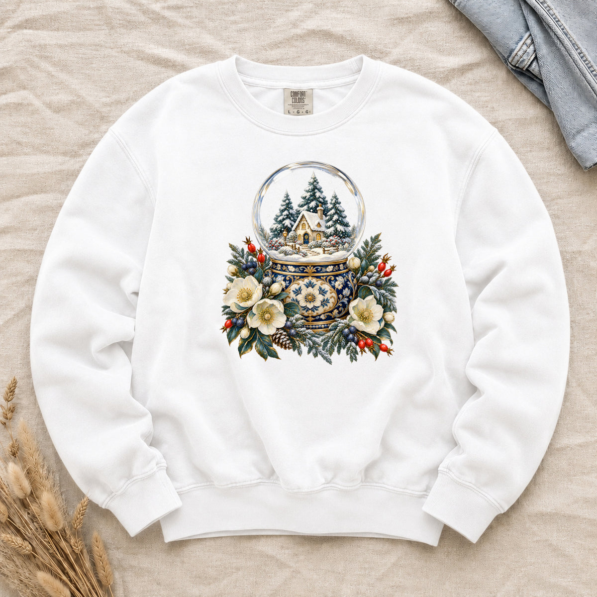 Snow Globe & Winter Florals Sweatshirt in White — a glass snow globe holding a snowy cottage and evergreens on an ornate navy-and-gold base, framed by cream blossoms, pinecones and red berries