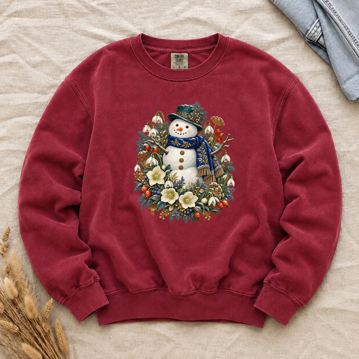Snowman & Winter Botanicals Sweatshirt in Crimson — a smiling snowman in a decorated dark hat and a deep blue scarf with golden pattern, among evergreen branches, red berries and large cream flowers