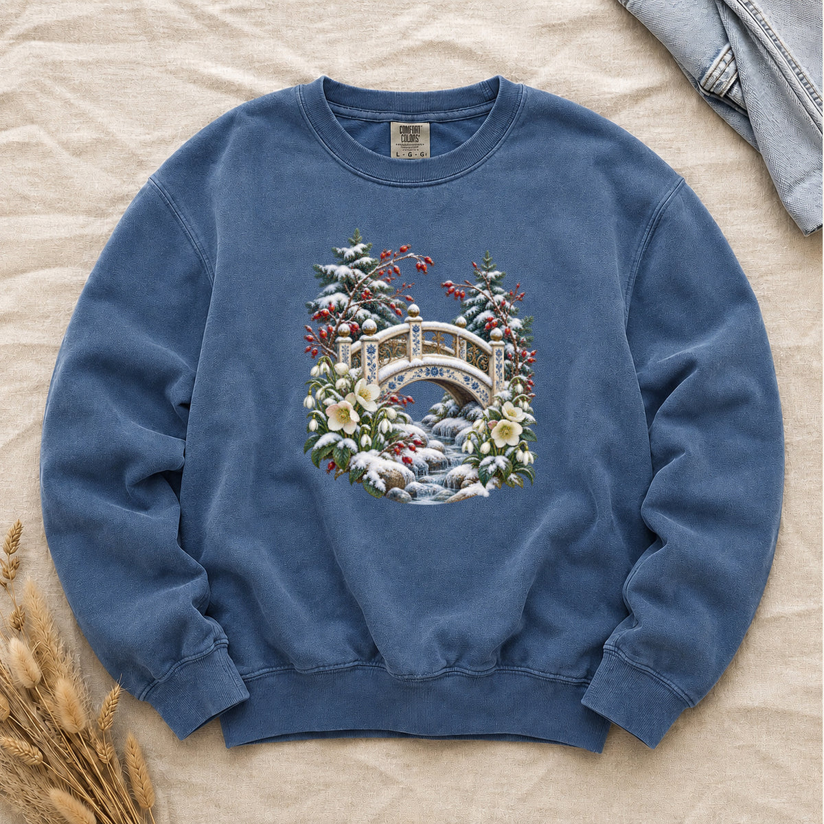 Snowy Bridge & Winter Botanicals Sweatshirt in Blue Jean — an ornate ivory footbridge with blue and golden details arching over a rocky stream, snow on its railings and on the evergreen around it, red berries and cream flowers framing the scene