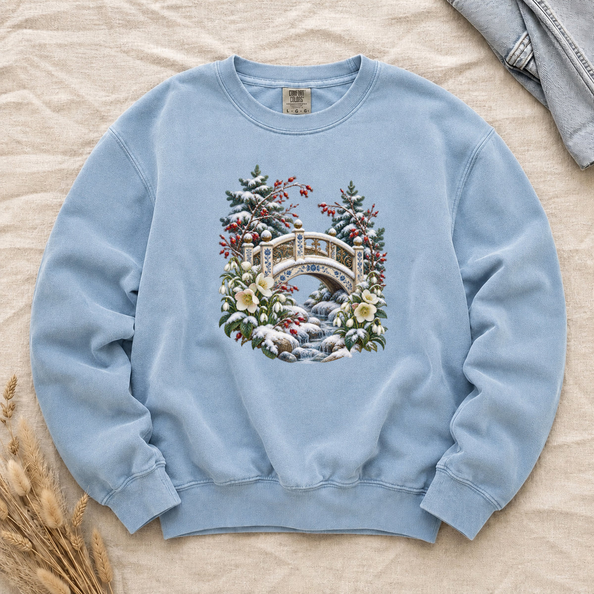 Snowy Bridge & Winter Botanicals Sweatshirt in Chambray — an ornate ivory footbridge with blue and golden details arching over a rocky stream, snow on its railings and on the evergreen around it, red berries and cream flowers framing the scene