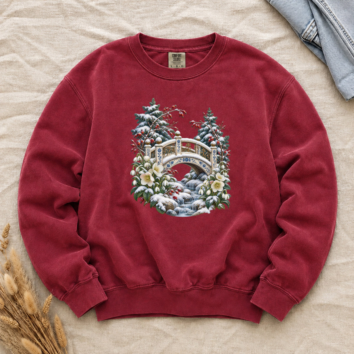 Snowy Bridge & Winter Botanicals Sweatshirt in Crimson — an ornate ivory footbridge with blue and golden details arching over a rocky stream, snow on its railings and on the evergreen around it, red berries and cream flowers framing the scene