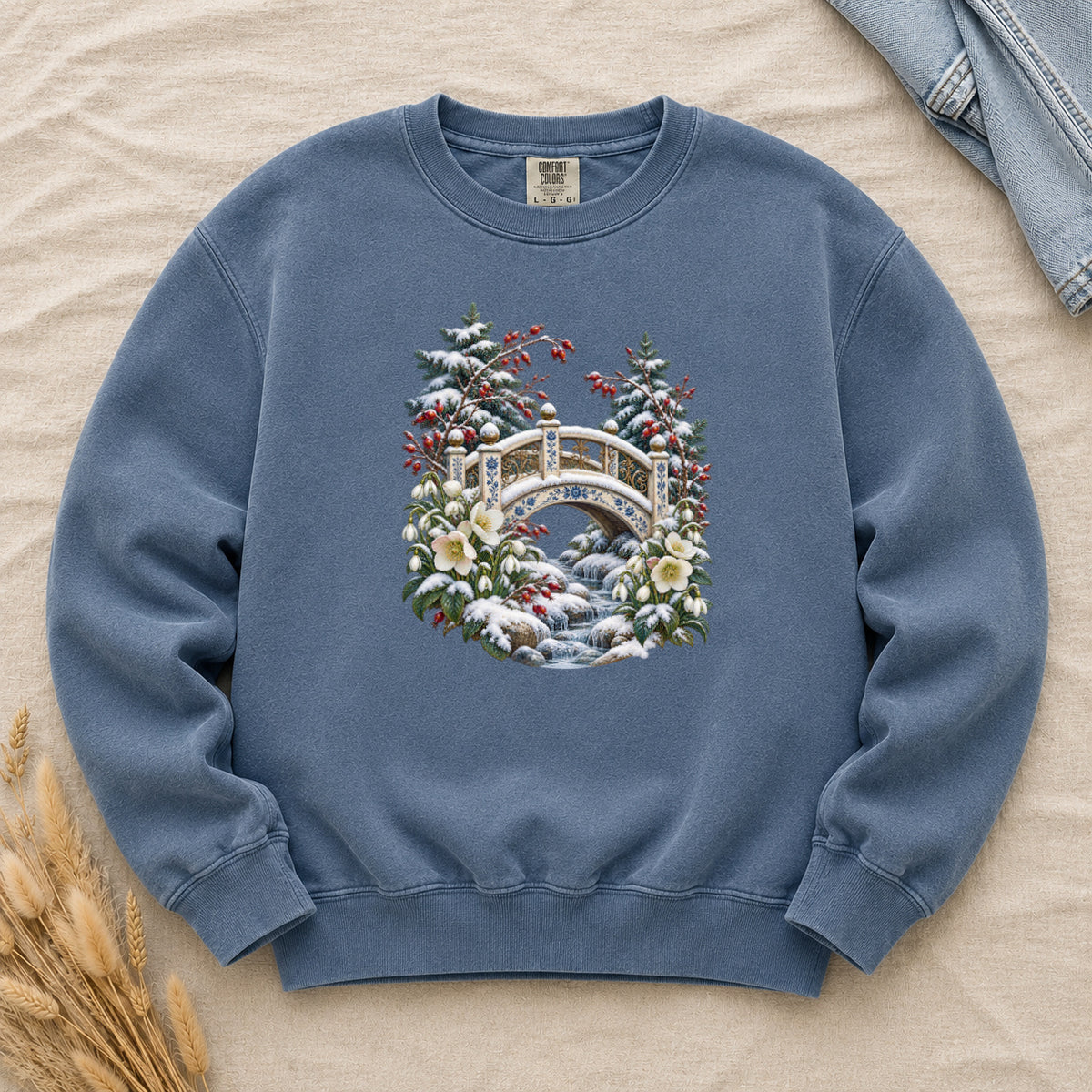 Snowy Bridge & Winter Botanicals Sweatshirt in Denim — an ornate ivory footbridge with blue and golden details arching over a rocky stream, snow on its railings and on the evergreen around it, red berries and cream flowers framing the scene