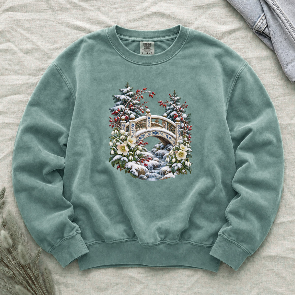 Snowy Bridge & Winter Botanicals Sweatshirt in Light Green — an ornate ivory footbridge with blue and golden details arching over a rocky stream, snow on its railings and on the evergreen around it, red berries and cream flowers framing the scene