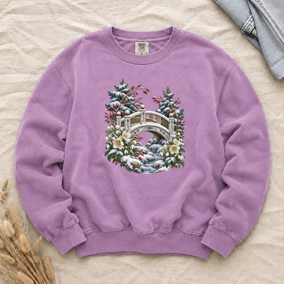 Snowy Bridge & Winter Botanicals Sweatshirt in Orchid — an ornate ivory footbridge with blue and golden details arching over a rocky stream, snow on its railings and on the evergreen around it, red berries and cream flowers framing the scene