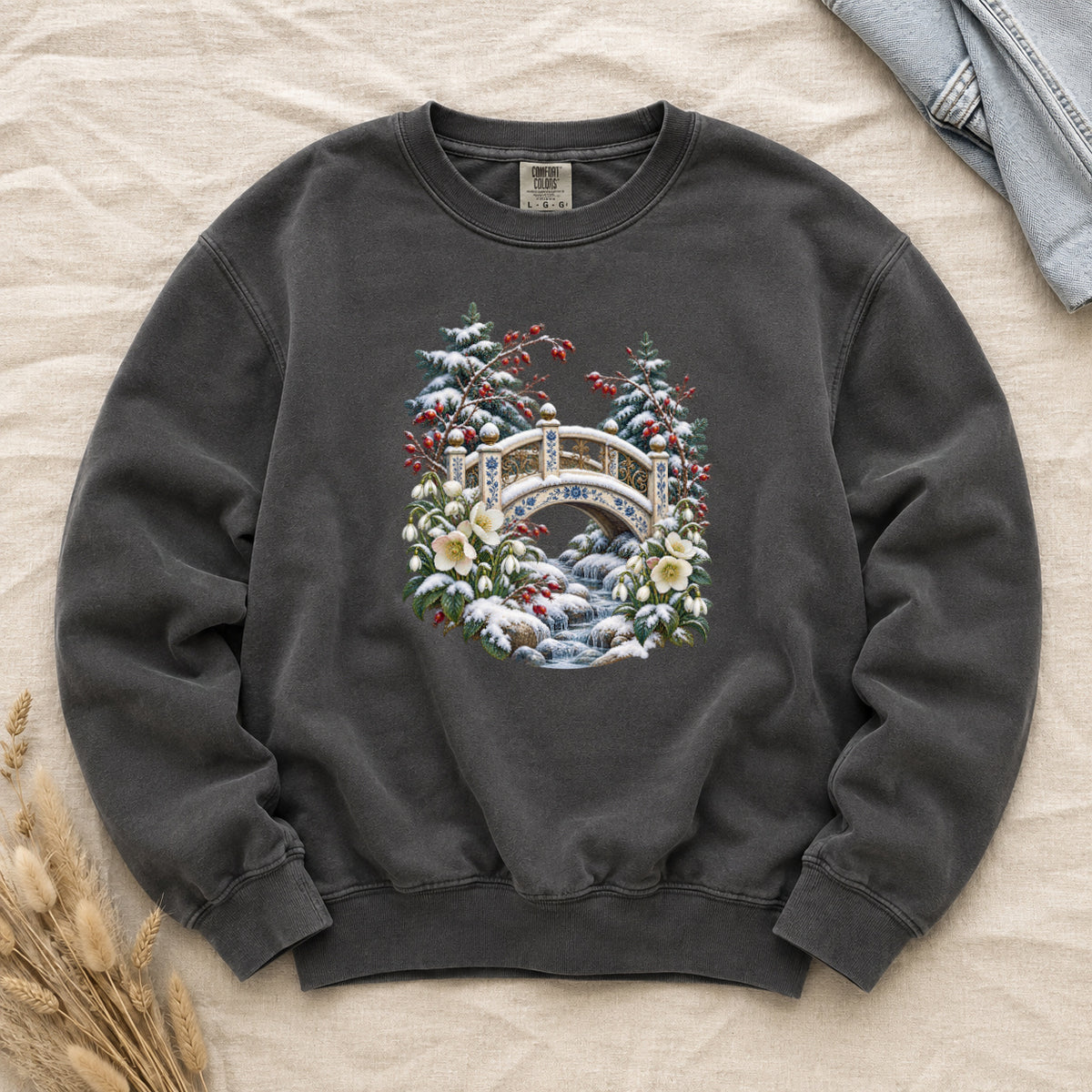 Snowy Bridge & Winter Botanicals Sweatshirt in Pepper — an ornate ivory footbridge with blue and golden details arching over a rocky stream, snow on its railings and on the evergreen around it, red berries and cream flowers framing the scene
