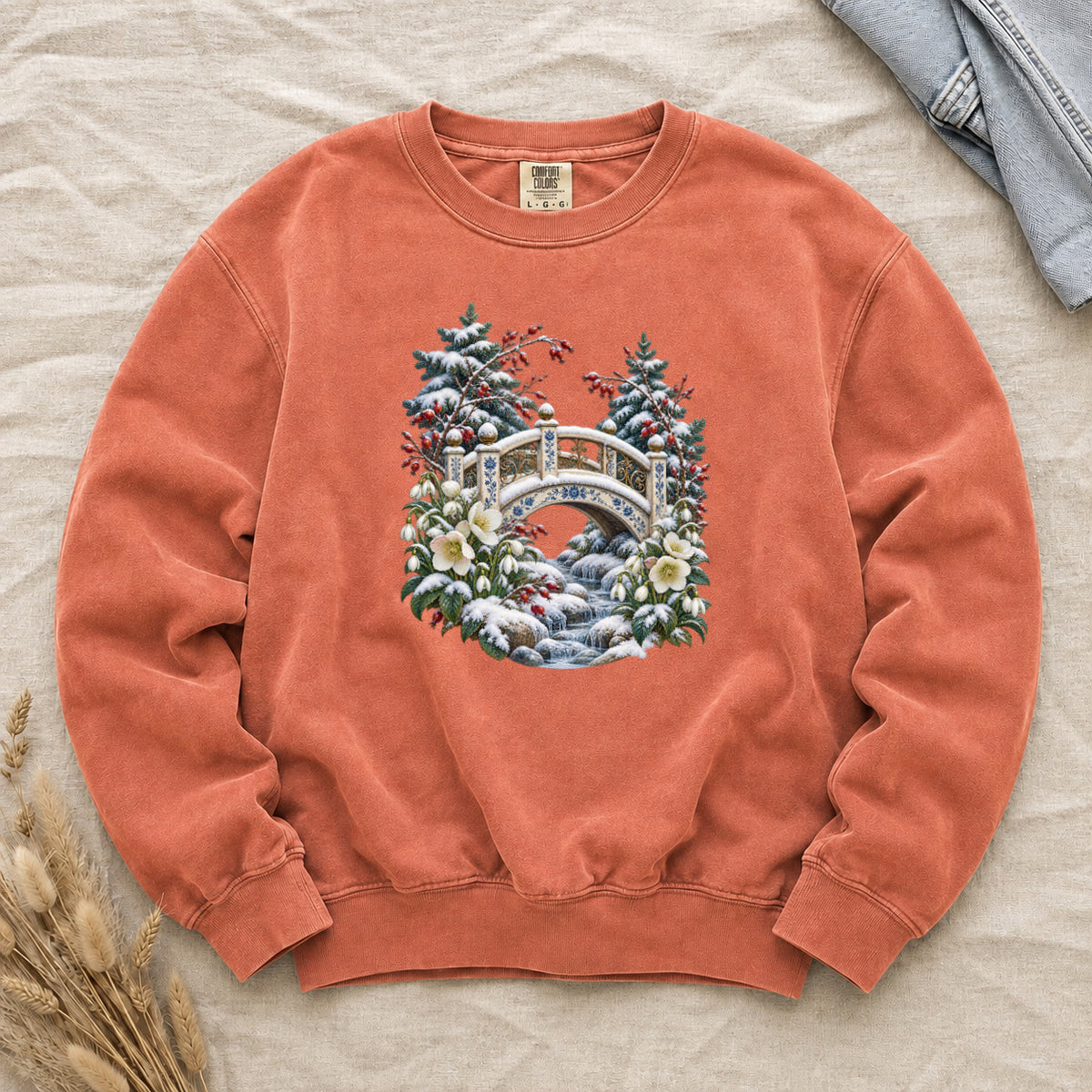 Snowy Bridge & Winter Botanicals Sweatshirt in Terracotta — an ornate ivory footbridge with blue and golden details arching over a rocky stream, snow on its railings and on the evergreen around it, red berries and cream flowers framing the scene