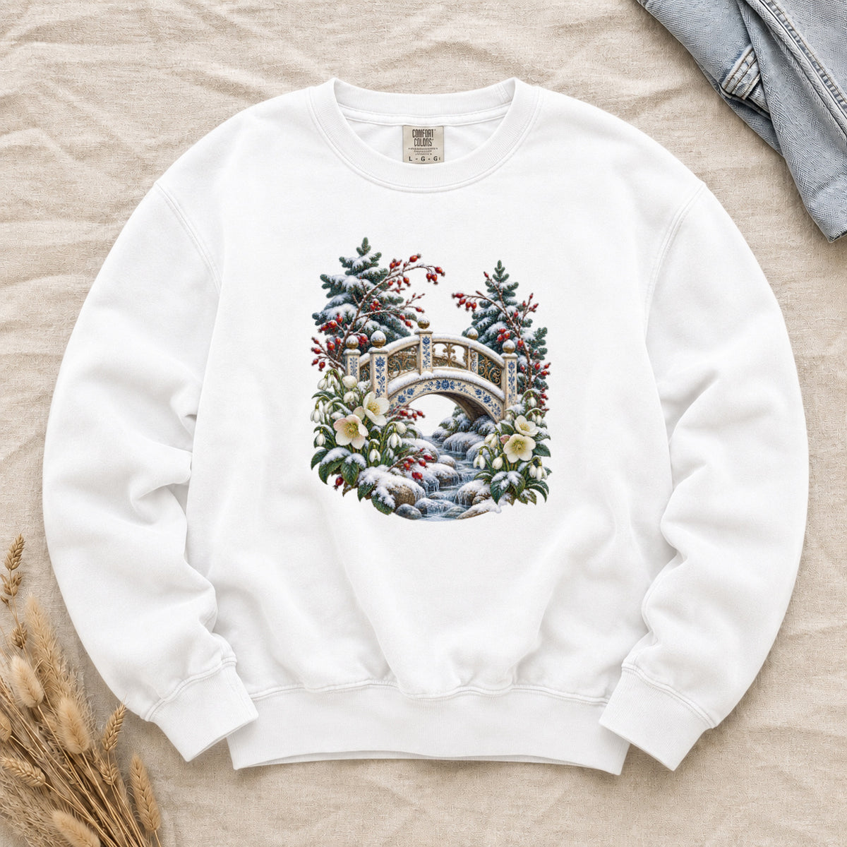 Snowy Bridge & Winter Botanicals Sweatshirt in White — an ornate ivory footbridge with blue and golden details arching over a rocky stream, snow on its railings and on the evergreen around it, red berries and cream flowers framing the scene