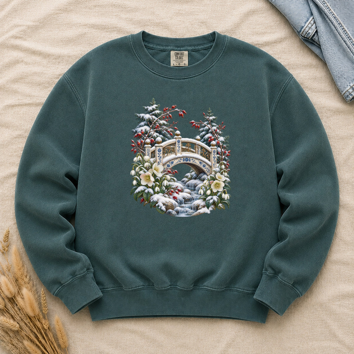 Snowy Bridge & Winter Botanicals Sweatshirt in Blue Spruce — an ornate ivory footbridge with blue and golden details arching over a rocky stream, snow on its railings and on the evergreen around it, red berries and cream flowers framing the scene