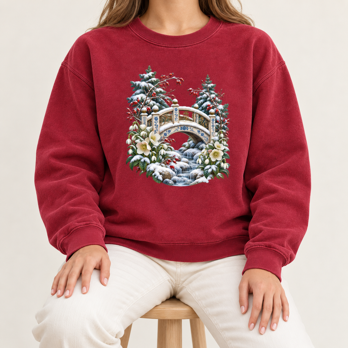 Snowy bridge and winter botanicals print on a 9.5 oz garment-dyed cotton-blend crewneck — an ornate ivory footbridge arching over a snowy rocky stream, framed by evergreen, red berries and cream flowers