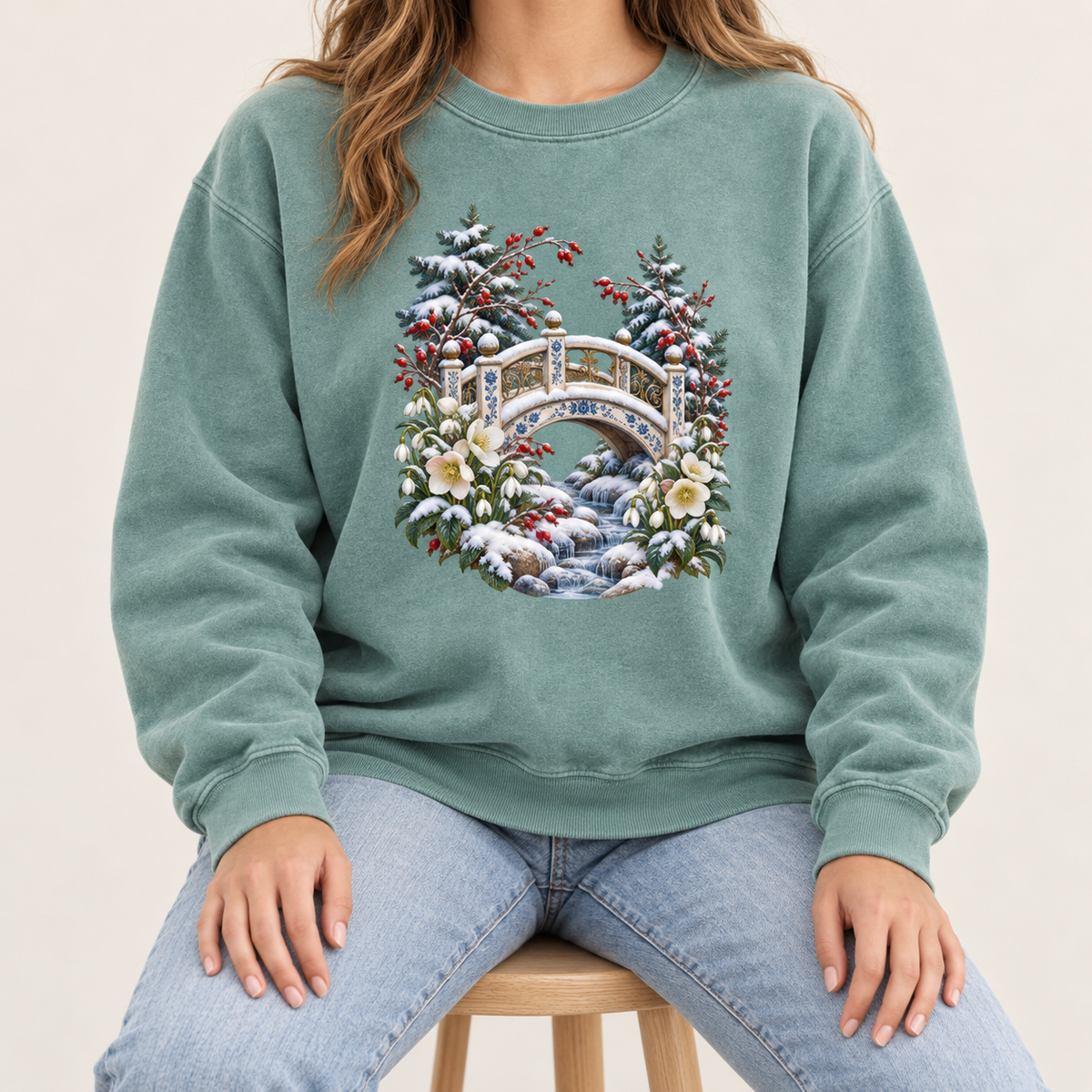 Snowy bridge design on a twill-taped, garment-dyed heavyweight crewneck — an ornate ivory footbridge arching over a snowy rocky stream, framed by evergreen, red berries and cream flowers