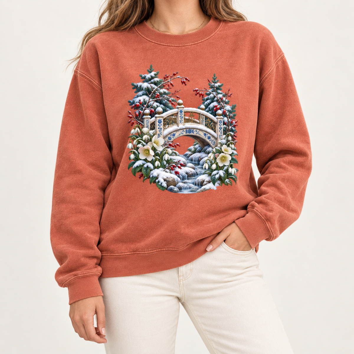 Snowy bridge winter crewneck, dyed after the garment is sewn with salt-free pigment dyes — an ornate ivory footbridge arching over a snowy rocky stream, framed by evergreen, red berries and cream flowers