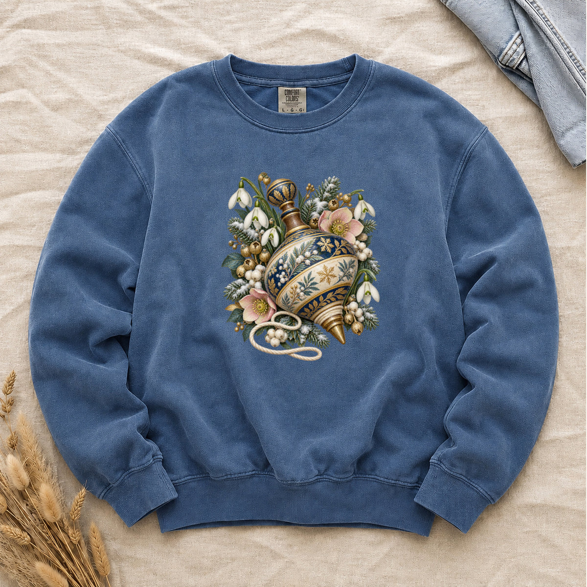 Vintage Spinning Top & Winter Florals Sweatshirt in Blue Jean — an ornate spinning top in deep blue, cream and gold, resting among evergreen sprigs, white snowdrops, pink flowers and pale berries, a loose cord curling beneath its tip