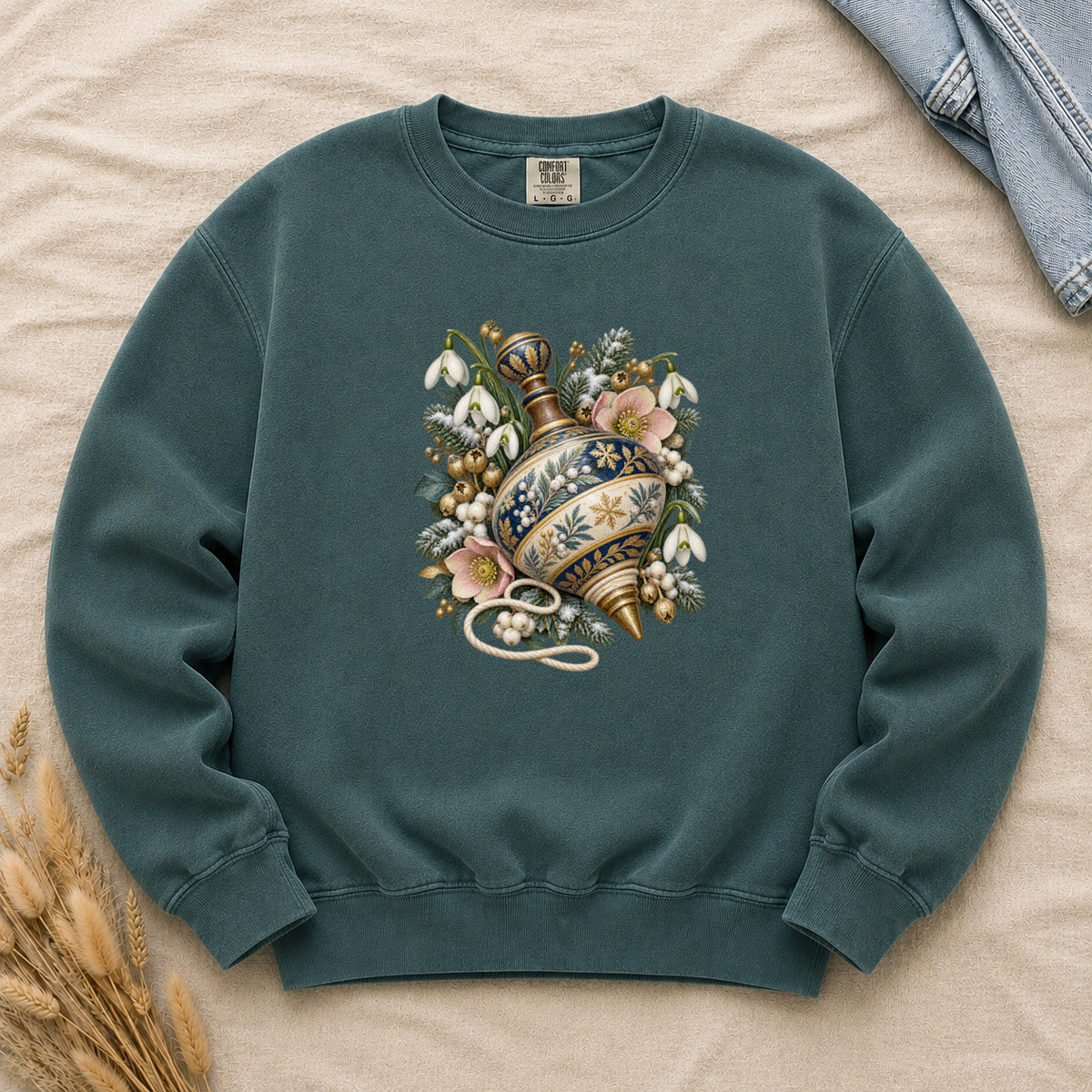 Vintage Spinning Top & Winter Florals Sweatshirt in Blue Spruce — an ornate spinning top in deep blue, cream and gold, resting among evergreen sprigs, white snowdrops, pink flowers and pale berries, a loose cord curling beneath its tip