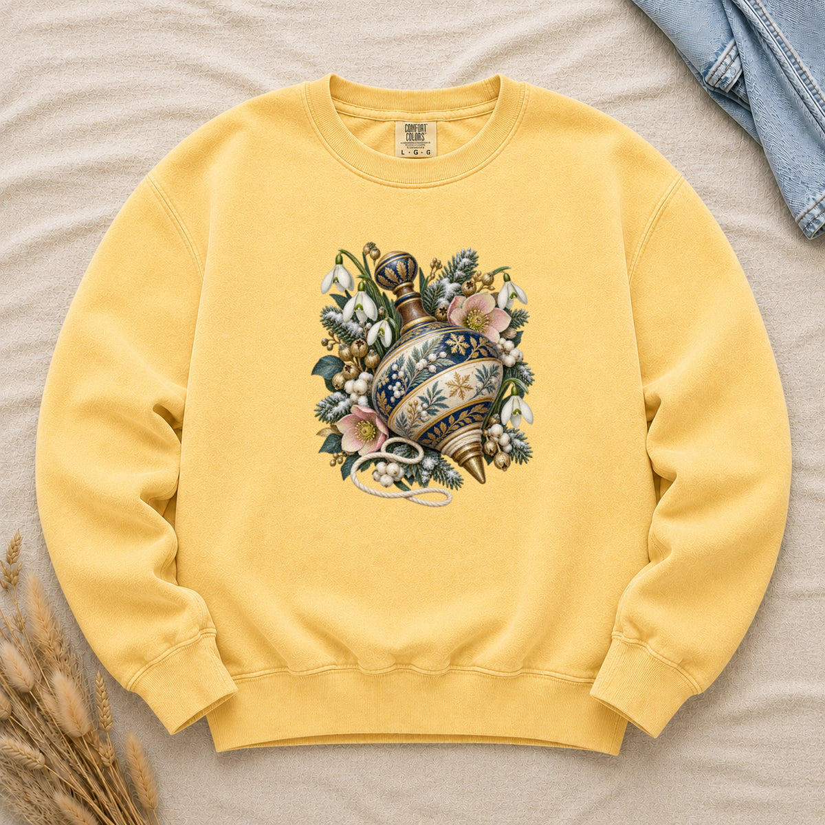 Vintage Spinning Top & Winter Florals Sweatshirt in Butter — an ornate spinning top in deep blue, cream and gold, resting among evergreen sprigs, white snowdrops, pink flowers and pale berries, a loose cord curling beneath its tip