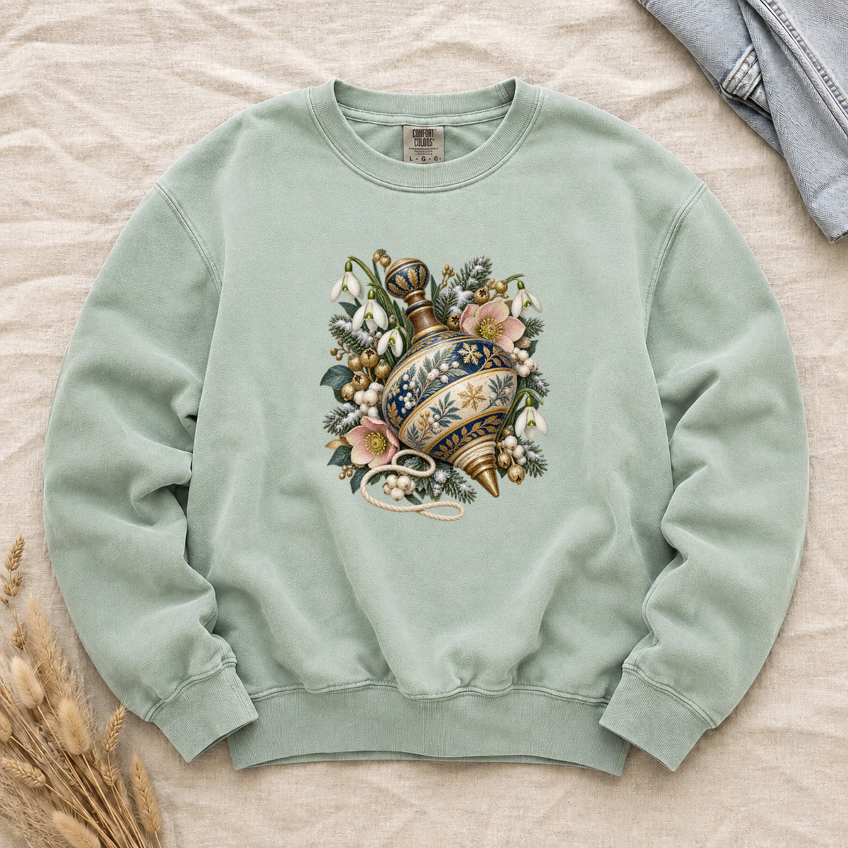 Vintage Spinning Top & Winter Florals Sweatshirt in Chalky Mint — an ornate spinning top in deep blue, cream and gold, resting among evergreen sprigs, white snowdrops, pink flowers and pale berries, a loose cord curling beneath its tip