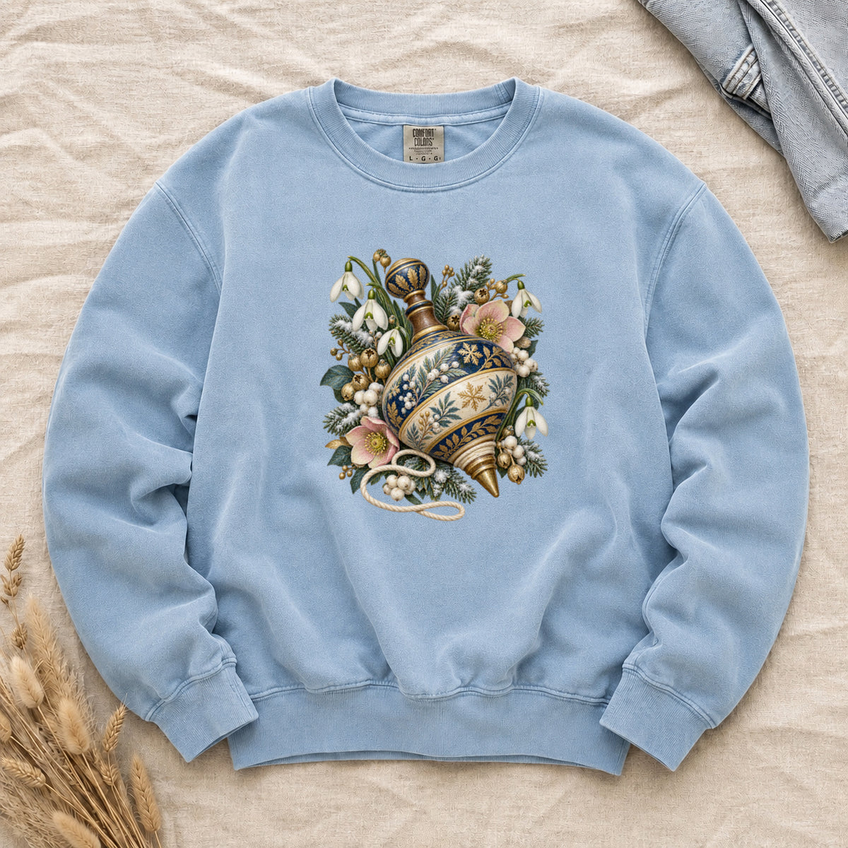 Vintage Spinning Top & Winter Florals Sweatshirt in Chambray — an ornate spinning top in deep blue, cream and gold, resting among evergreen sprigs, white snowdrops, pink flowers and pale berries, a loose cord curling beneath its tip