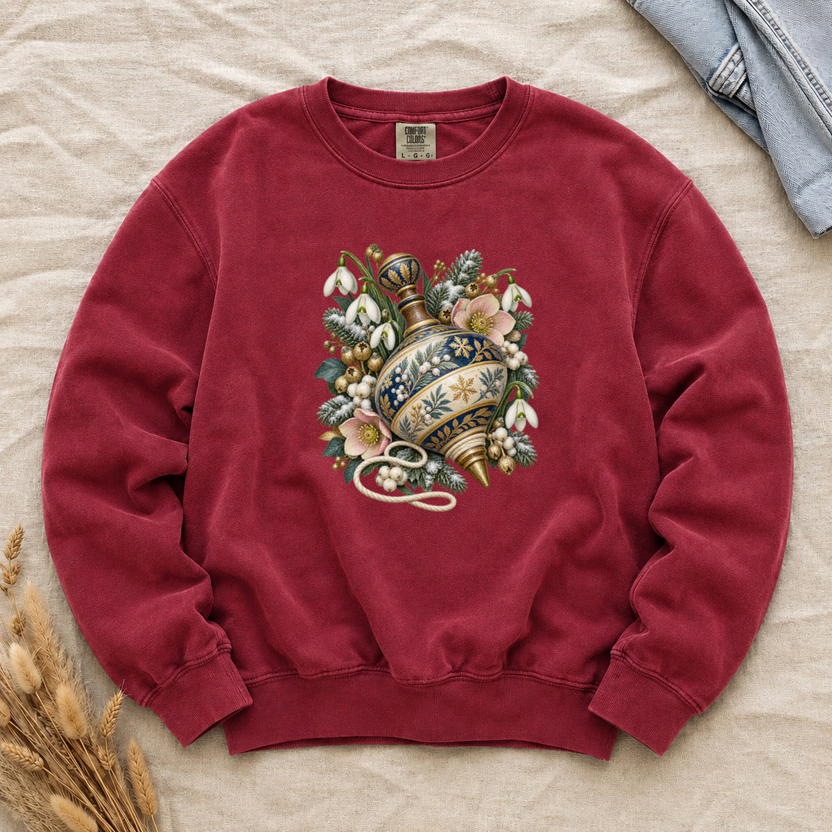Vintage Spinning Top & Winter Florals Sweatshirt in Crimson — an ornate spinning top in deep blue, cream and gold, resting among evergreen sprigs, white snowdrops, pink flowers and pale berries, a loose cord curling beneath its tip