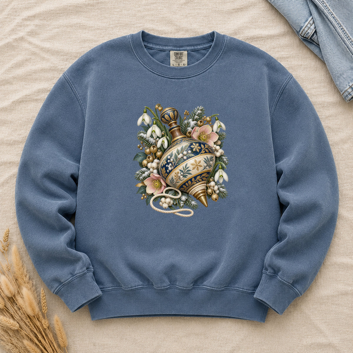 Vintage Spinning Top & Winter Florals Sweatshirt in Denim — an ornate spinning top in deep blue, cream and gold, resting among evergreen sprigs, white snowdrops, pink flowers and pale berries, a loose cord curling beneath its tip