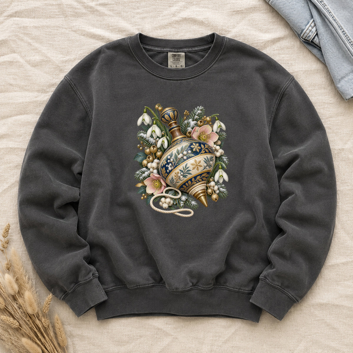 Vintage Spinning Top & Winter Florals Sweatshirt in Pepper — an ornate spinning top in deep blue, cream and gold, resting among evergreen sprigs, white snowdrops, pink flowers and pale berries, a loose cord curling beneath its tip