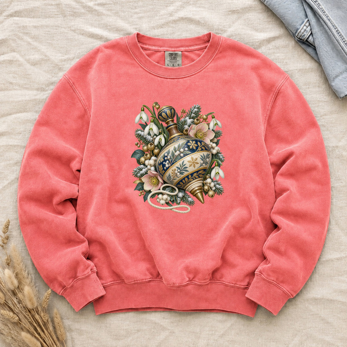 Vintage Spinning Top & Winter Florals Sweatshirt in Watermelon — an ornate spinning top in deep blue, cream and gold, resting among evergreen sprigs, white snowdrops, pink flowers and pale berries, a loose cord curling beneath its tip