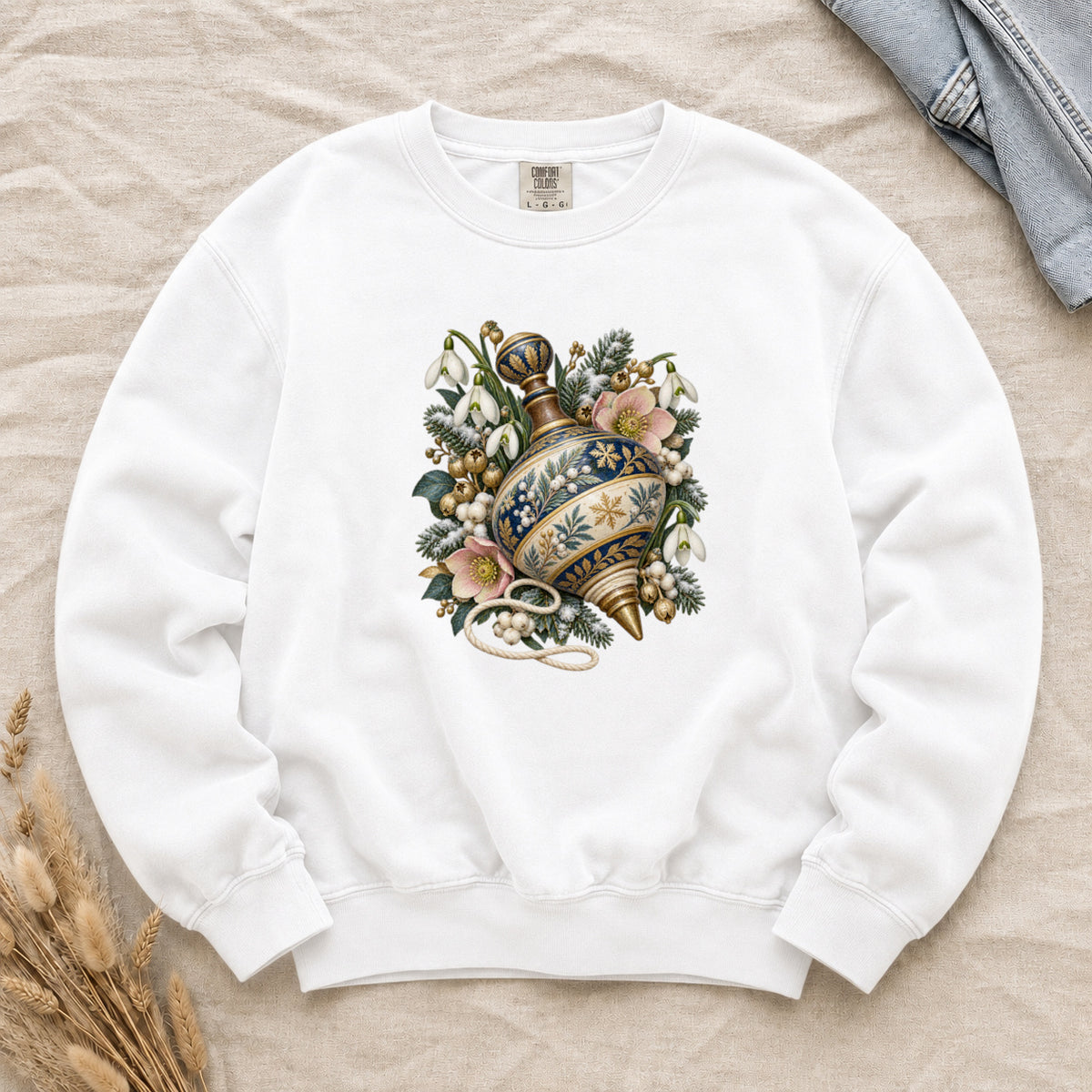 Vintage Spinning Top & Winter Florals Sweatshirt in White — an ornate spinning top in deep blue, cream and gold, resting among evergreen sprigs, white snowdrops, pink flowers and pale berries, a loose cord curling beneath its tip
