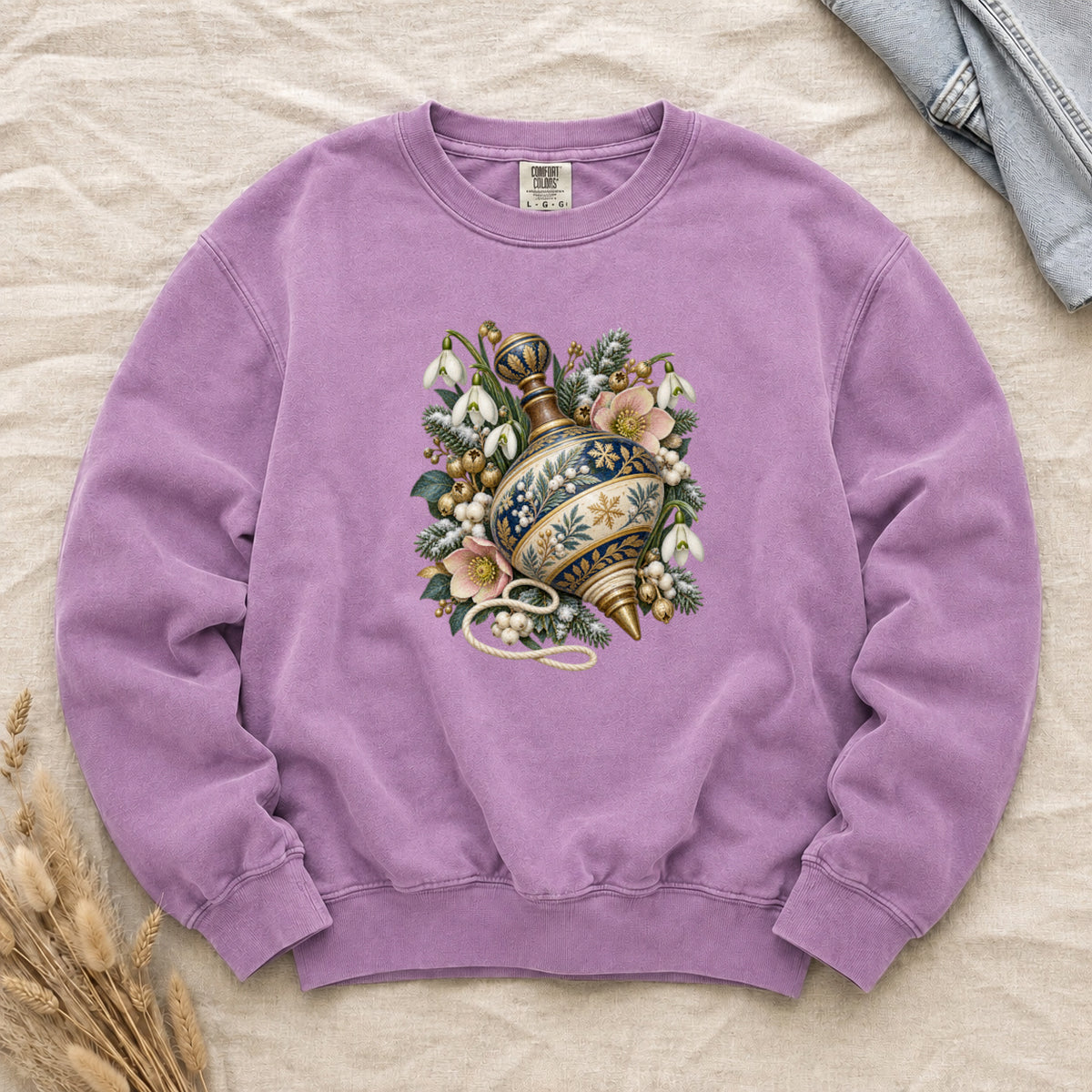 Vintage Spinning Top & Winter Florals Sweatshirt in Orchid — an ornate spinning top in deep blue, cream and gold, resting among evergreen sprigs, white snowdrops, pink flowers and pale berries, a loose cord curling beneath its tip