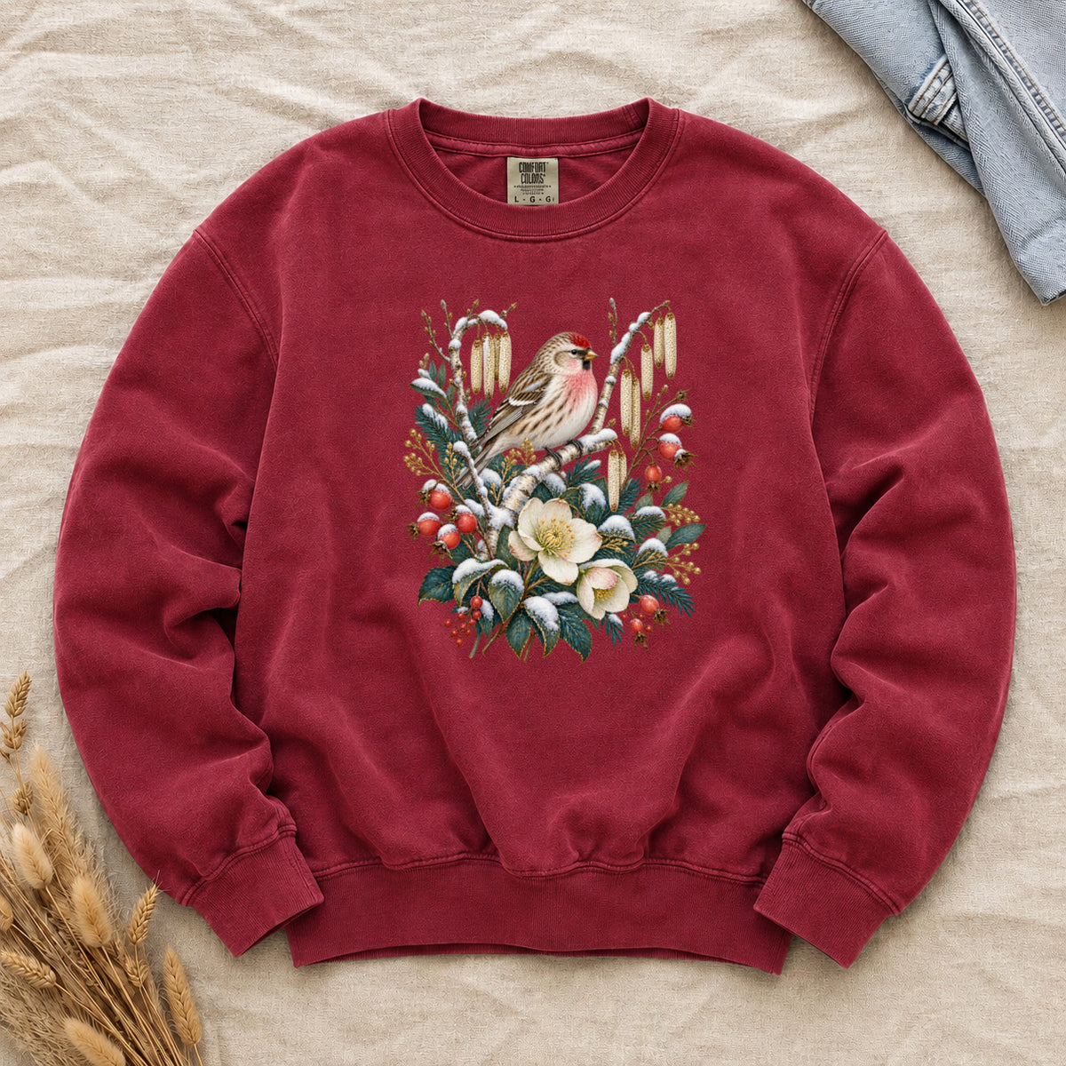 Winter Finch & Botanical Sweatshirt in Crimson — a small brown finch with rosy red markings perched among branches hung with golden catkins, above snow-dusted evergreen, red berries and cream flowers