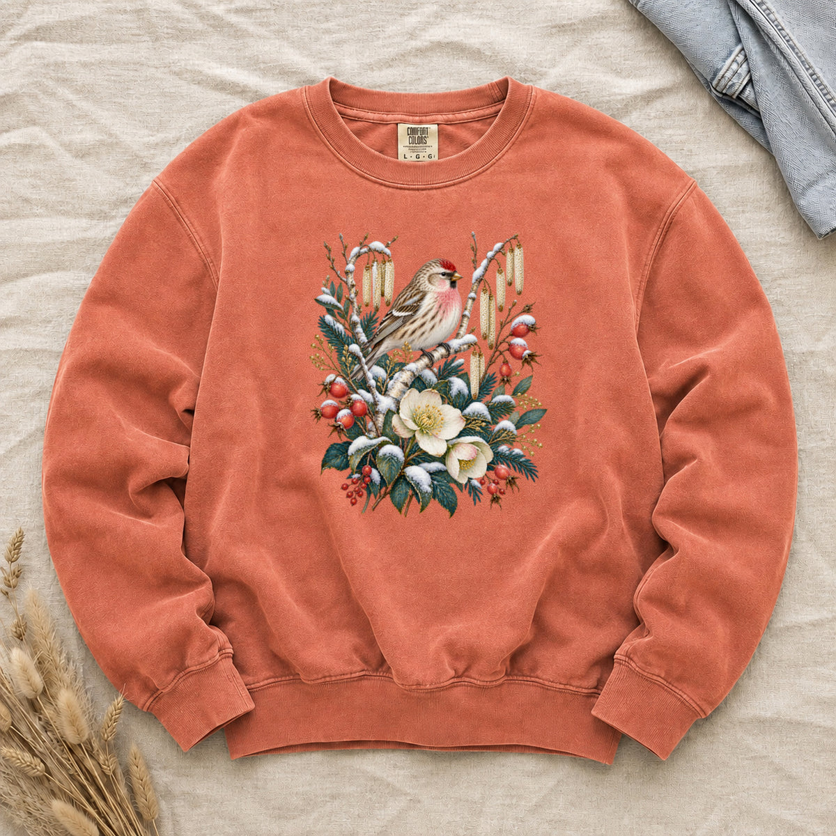 Winter Finch & Botanical Sweatshirt in Terracotta — a small brown finch with rosy red markings perched among branches hung with golden catkins, above snow-dusted evergreen, red berries and cream flowers