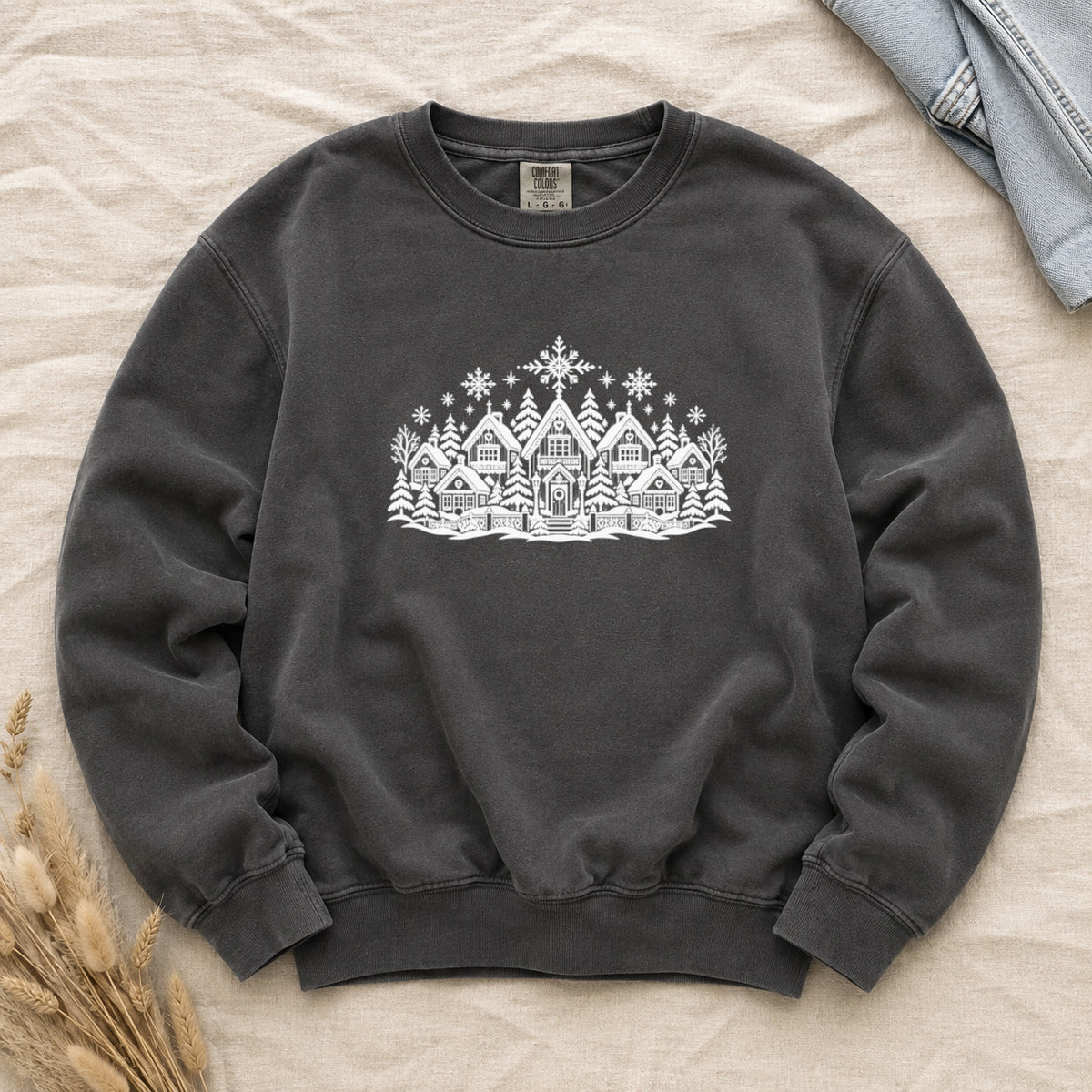 Winter Village White Line Art Sweatshirt in Pepper — a row of steep-roofed village houses with snow on the eaves, pines between them and a large snowflake above, drawn in white line art on a deep charcoal garment-dyed Comfort Colors crewneck