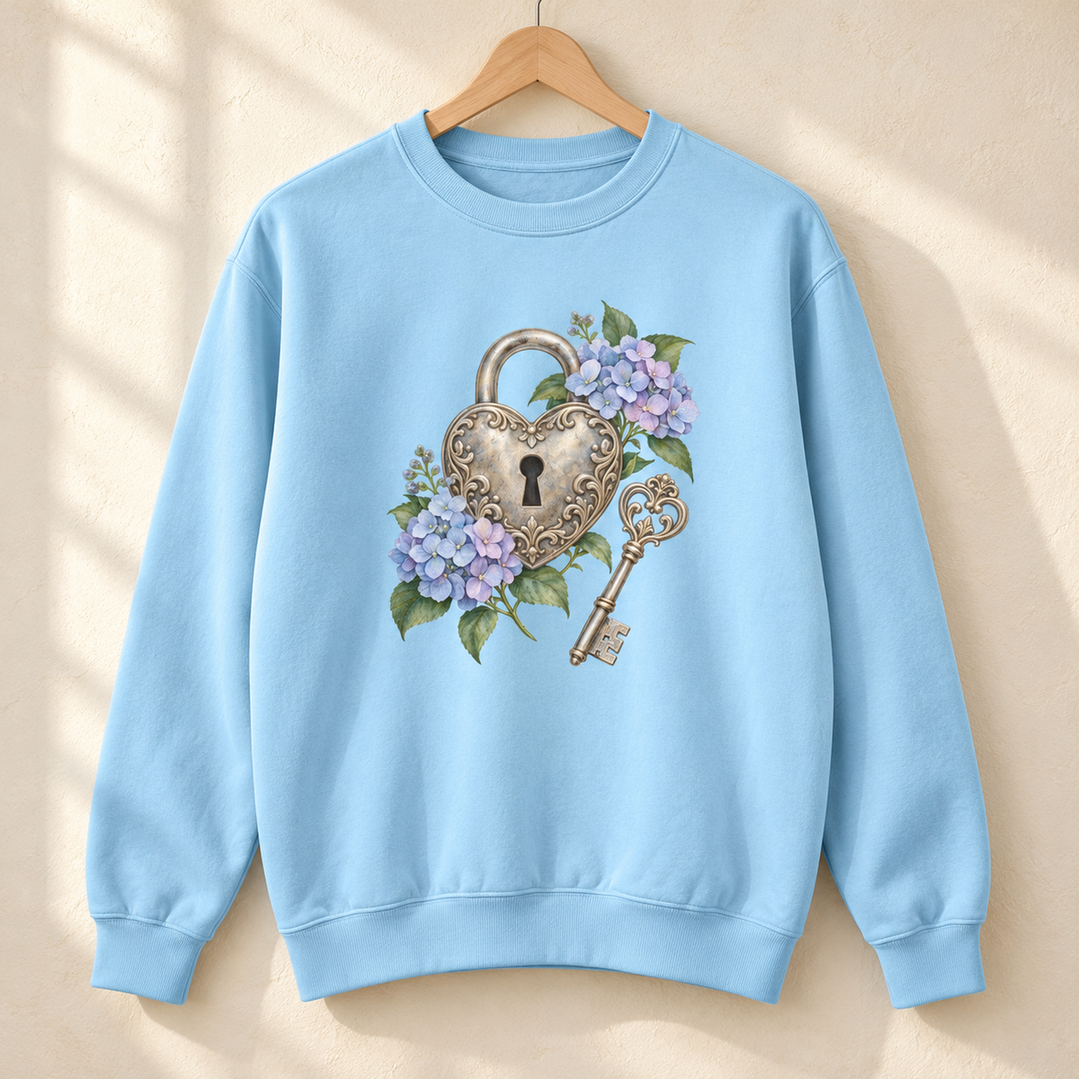 Detailed watercolor of a silver filigree heart padlock and skeleton key among blue and purple hydrangeas, printed large and centered on the front of a Chambray light blue 9.5 oz garment-dyed Comfort Colors 1566 sweatshirt, shown on a hanger