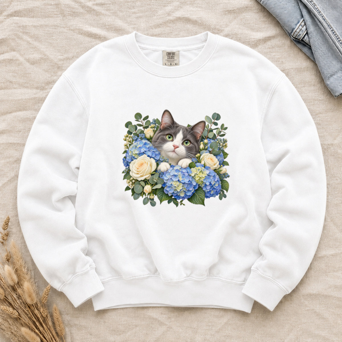 Tuxedo Cat and Blue Hydrangeas Sweatshirt in White — a grey-and-white tuxedo-pattern cat looking out from blue hydrangeas, cream roses and silver-green eucalyptus, printed large and centered on a garment-dyed Comfort Colors crewneck